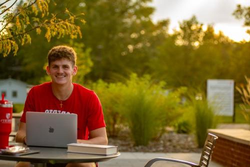 A young male student sits outside in front of a laptop and smiles.